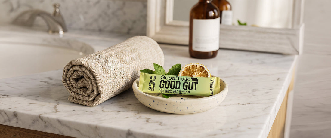 A bright bathroom counter in soft morning light, a rolled linen towel and a small ceramic dish holding a GoodGut sachet.