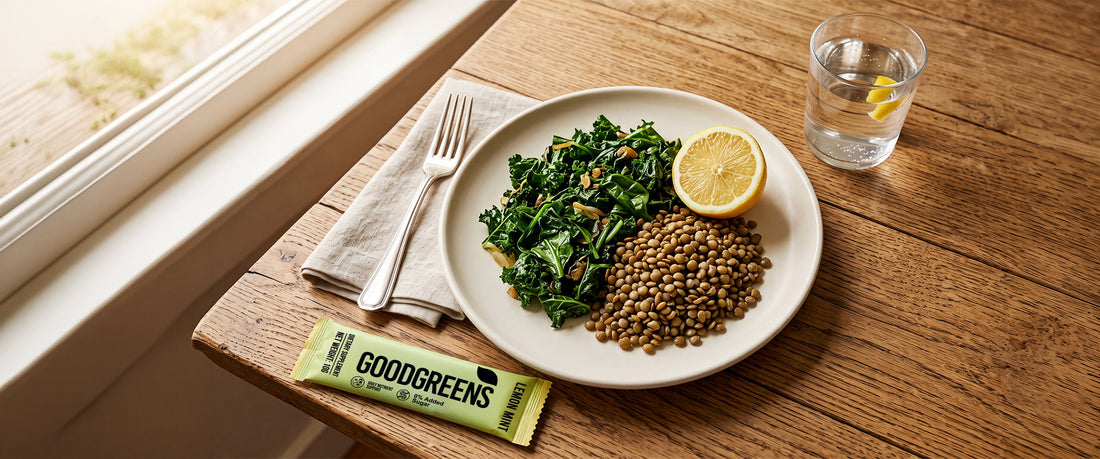 An overhead shot of a plate with dark leafy greens, lentils, and half a lemon in warm daylight, a glass of water and a GoodGreens sachet at the edge.