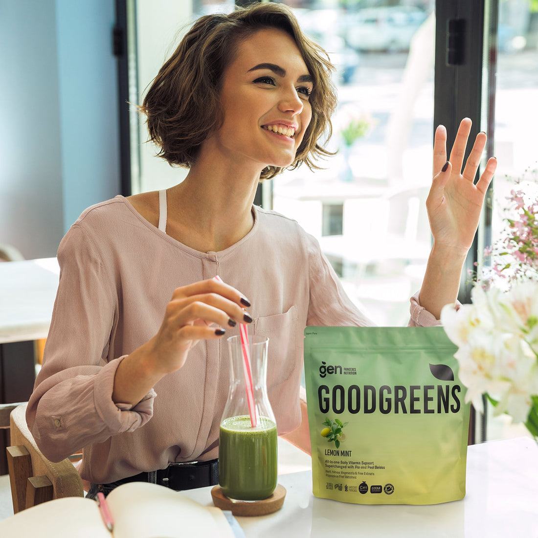 Smiling woman drinking GoodGreens Lemon Mint greens powder mixed in water, with the pack on the table