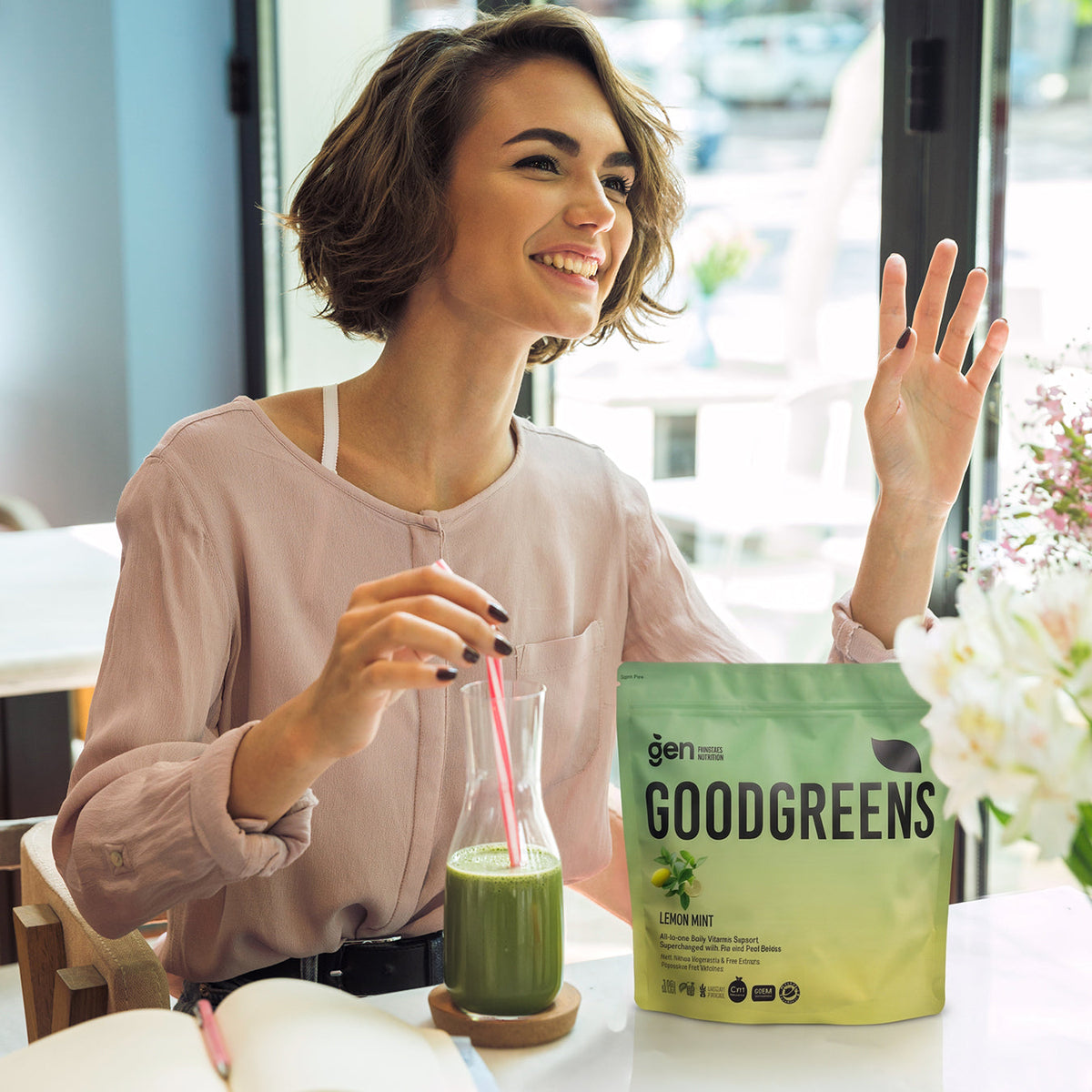Smiling woman drinking GoodGreens Lemon Mint greens powder mixed in water, with the pack on the table