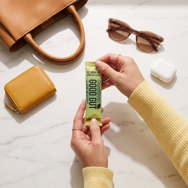 Hands holding a GoodGut sachet next to a handbag and sunglasses