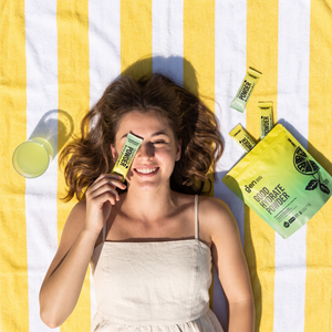 Smiling woman holding a GoodHydrate Lemon Mint sachet, packs beside her
